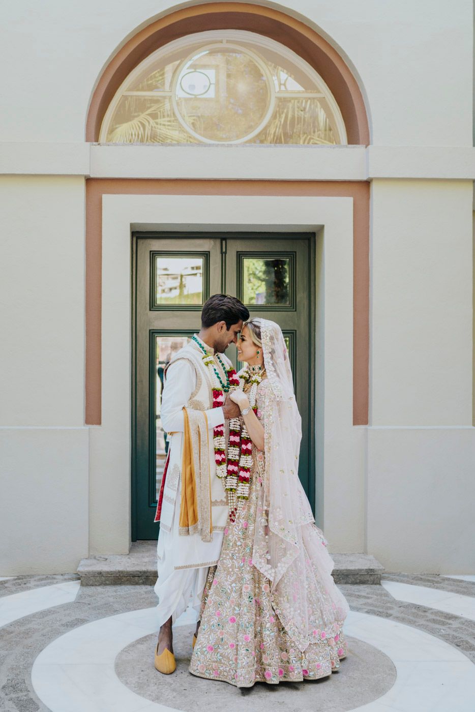 A couple in traditional Indian wedding attire stand in an intimate moment beneath an arched doorway.