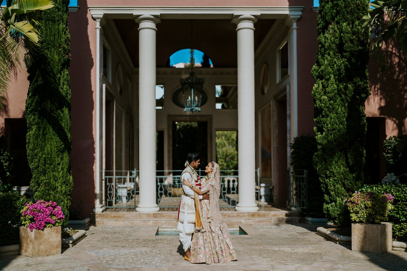 Newlyweds in elegant attire pose beneath classic columns of a grand entrance.