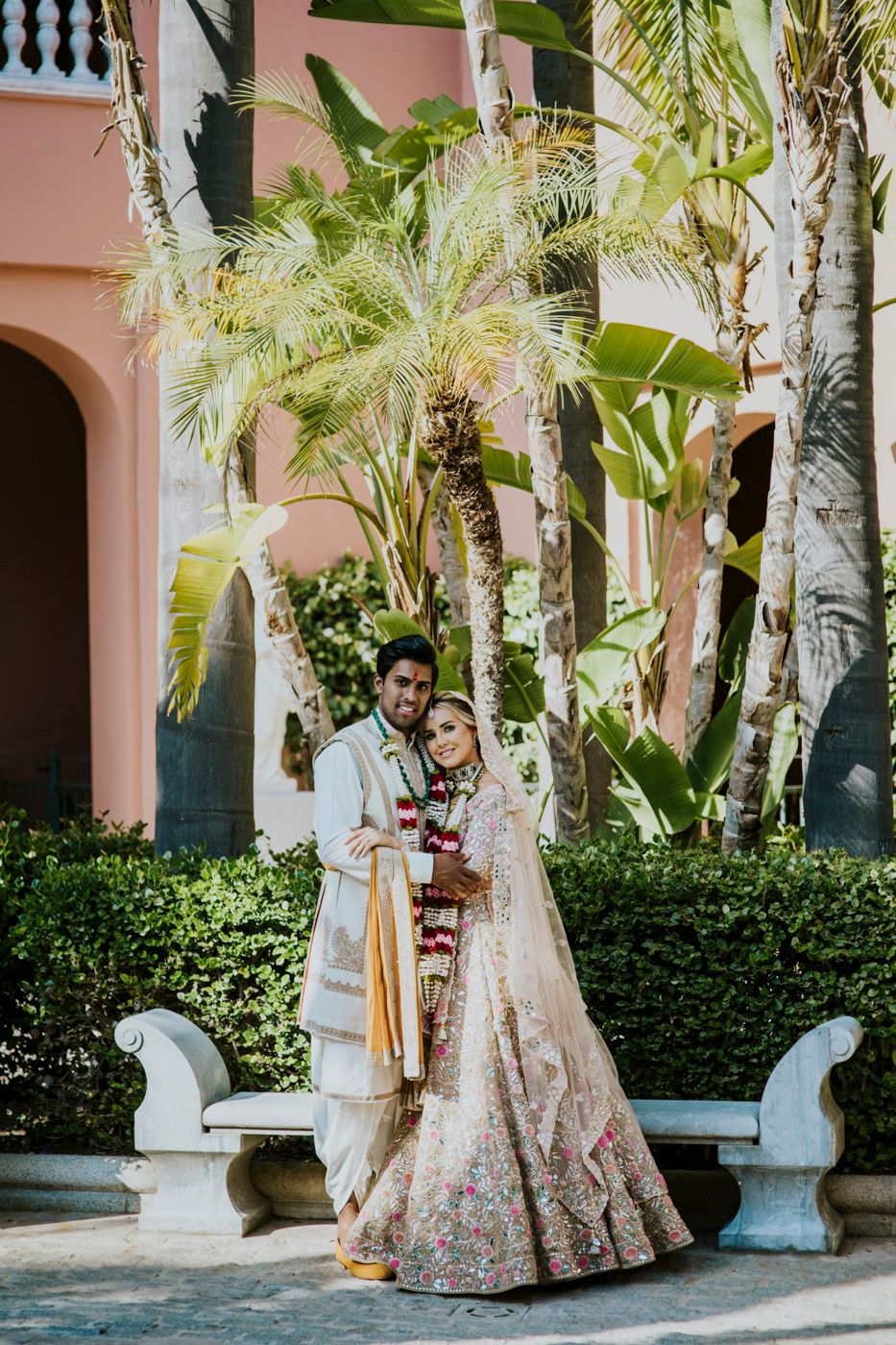 A couple in traditional Indian wedding attire poses together in front of tropical palm trees and a pink building.