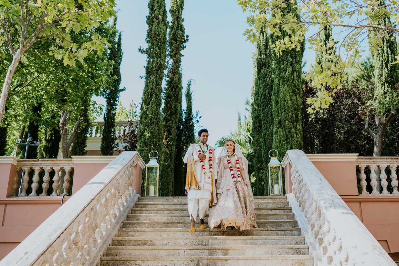 A couple descends an elegant stone staircase flanked by tall cypress trees in a Mediterranean style garden setting.