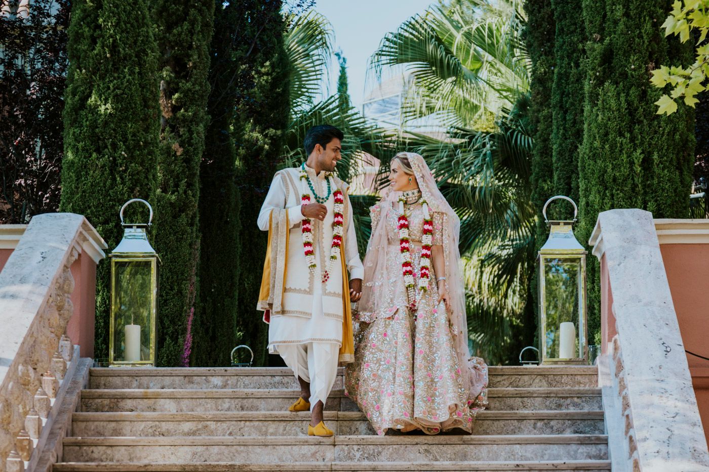 Couple in traditional Indian wedding outfits descending stone steps with decorative lanterns.