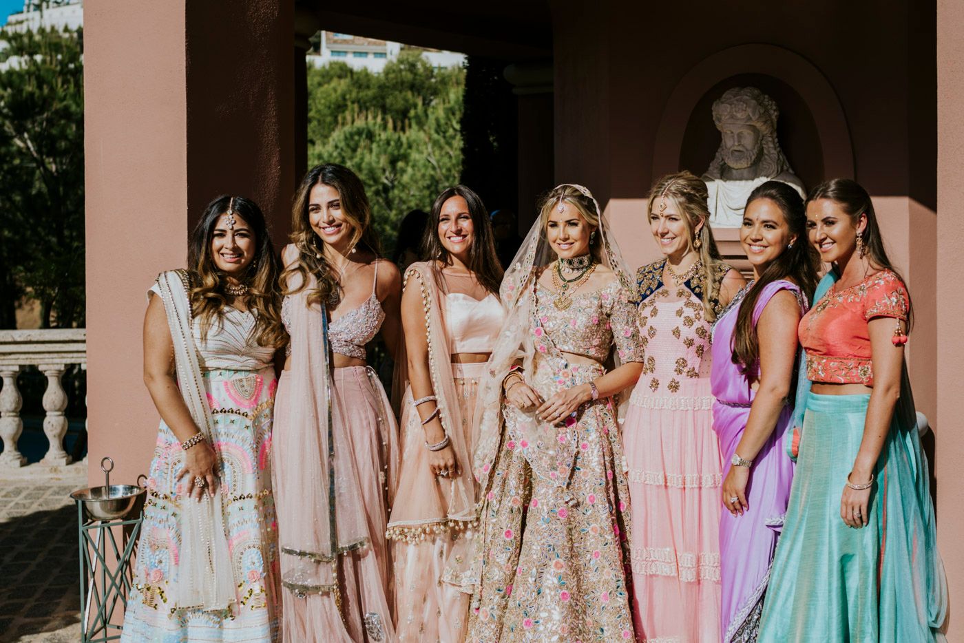 Wedding party in pastel dresses poses together on ornate balcony during outdoor celebration.