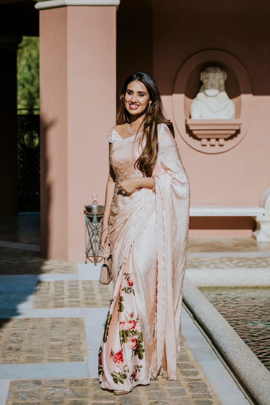 A woman wearing an elegant floral white and pink saree poses gracefully in front of a pink building with architectural details.