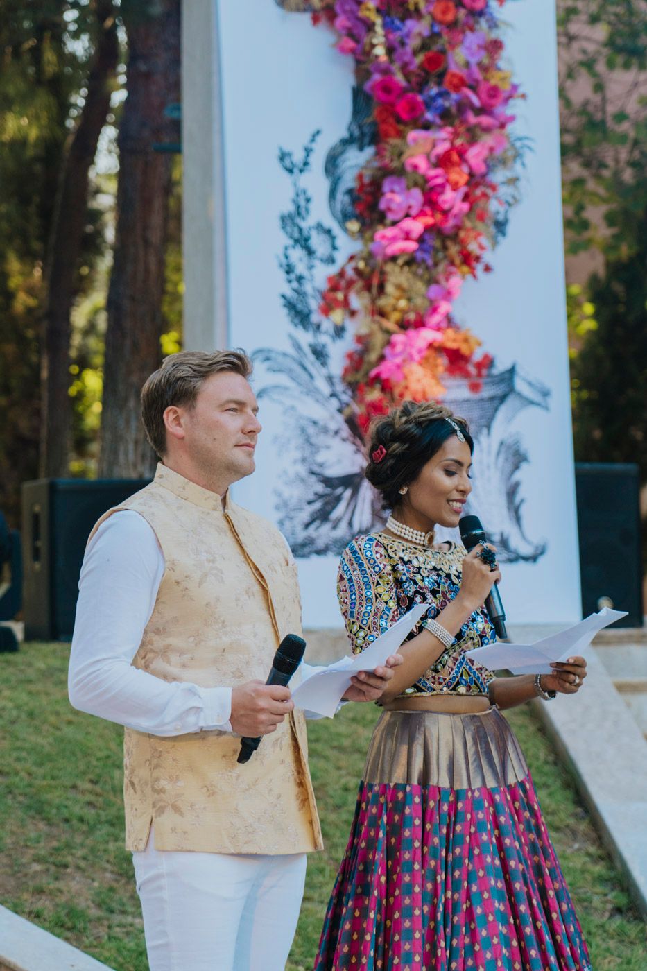 Two event speakers hold microphones in front of colorful floral backdrop during outdoor celebration.