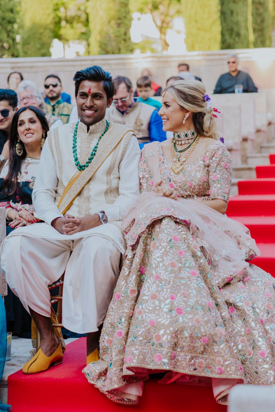 A couple in traditional South Asian wedding attire sits together on red carpeted stairs during a celebration.
