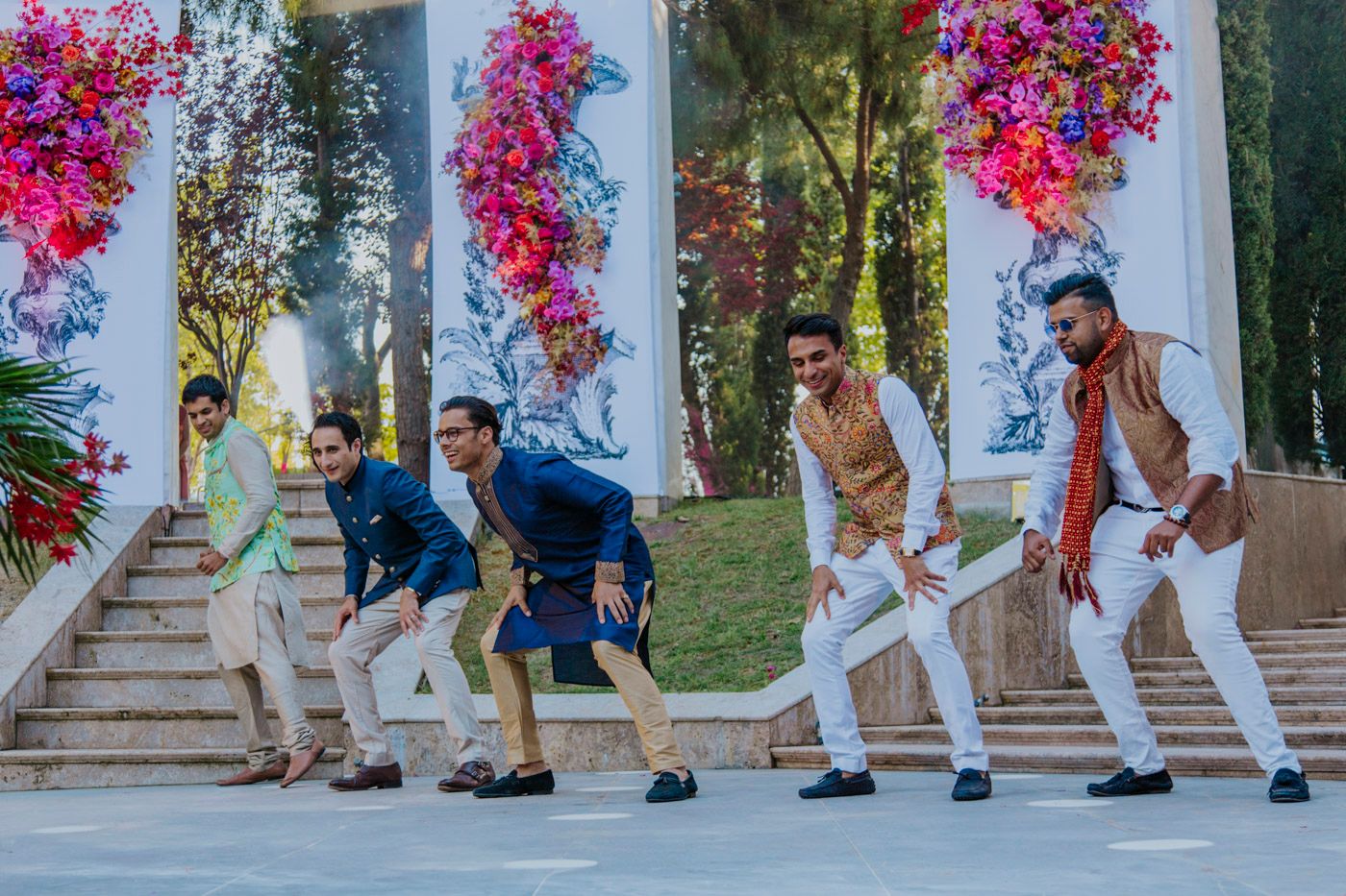 A group of groomsmen pose in a fun dance formation on stairs decorated with vibrant pink bougainvillea flowers.