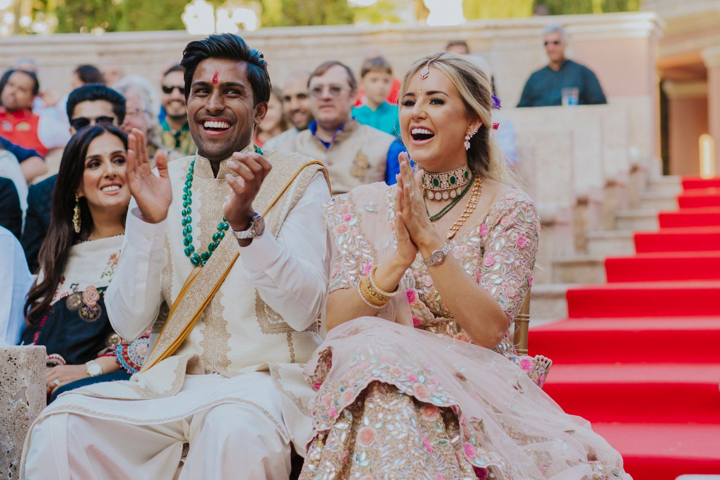 A couple in traditional South Asian wedding attire laugh joyfully while sitting together during their outdoor ceremony.