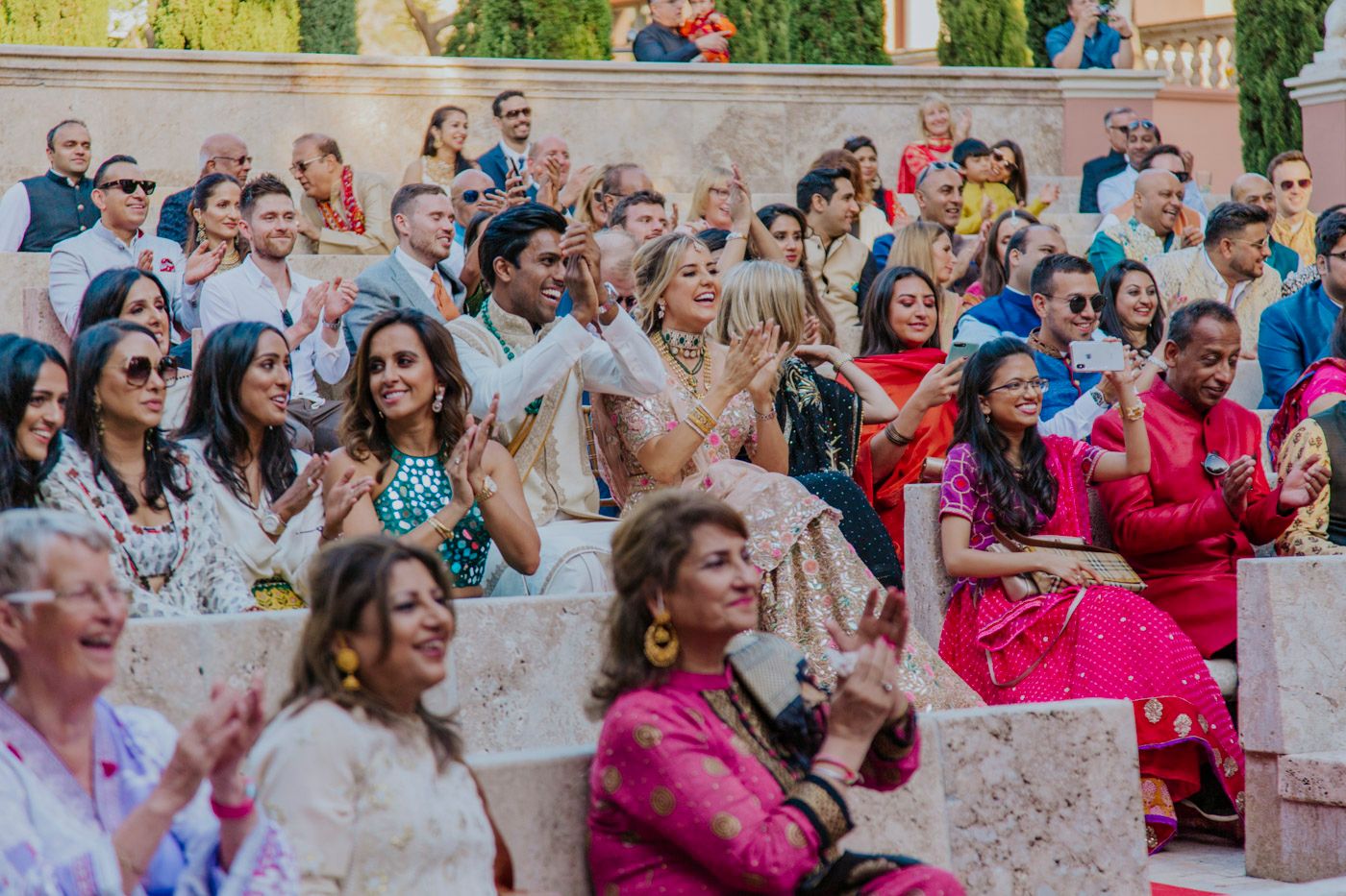 A colorful audience wearing traditional and modern attire claps and celebrates at an outdoor venue with bright floral decor.