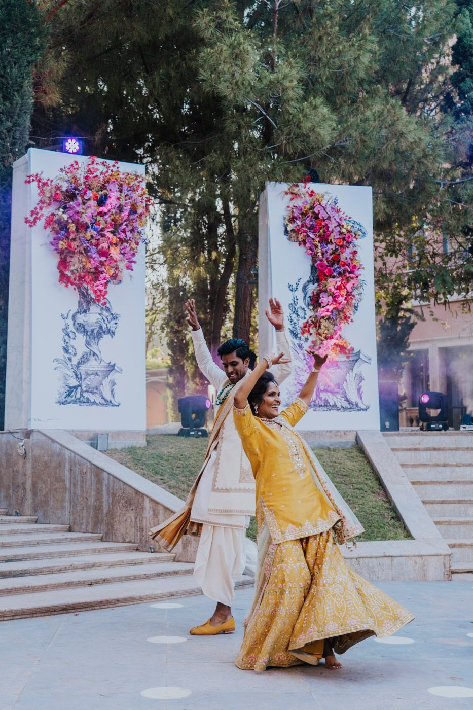 A couple in vibrant yellow traditional Indian attire dance joyfully in front of decorative floral wall panels at an outdoor venue.