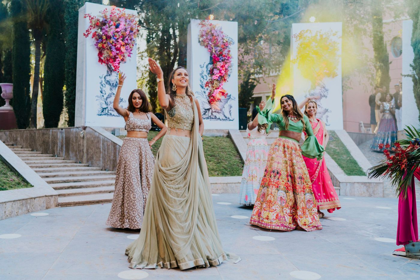 Elegant group in formal gowns gather at an outdoor event venue with pink floral decorations and gold accents.