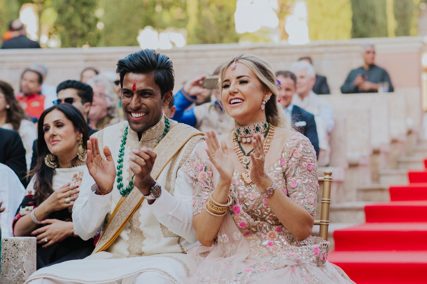 A couple at an outdoor ceremony clapping and smiling while seated on ornate chairs near red carpeted steps.