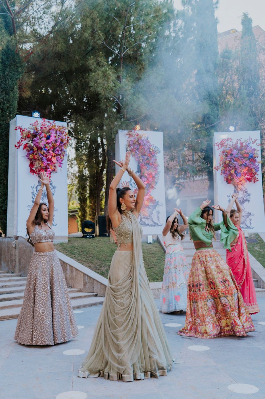 A festive celebration with dancers in traditional lehengas holding colorful smoke flares during an outdoor event.