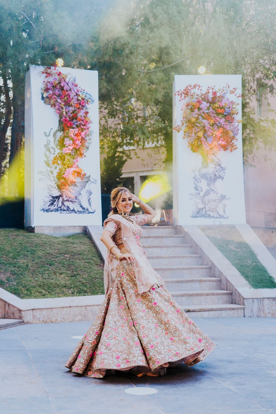 Dancing woman in ornate pink and gold lehenga twirls in front of colorful floral art installations on white pillars.