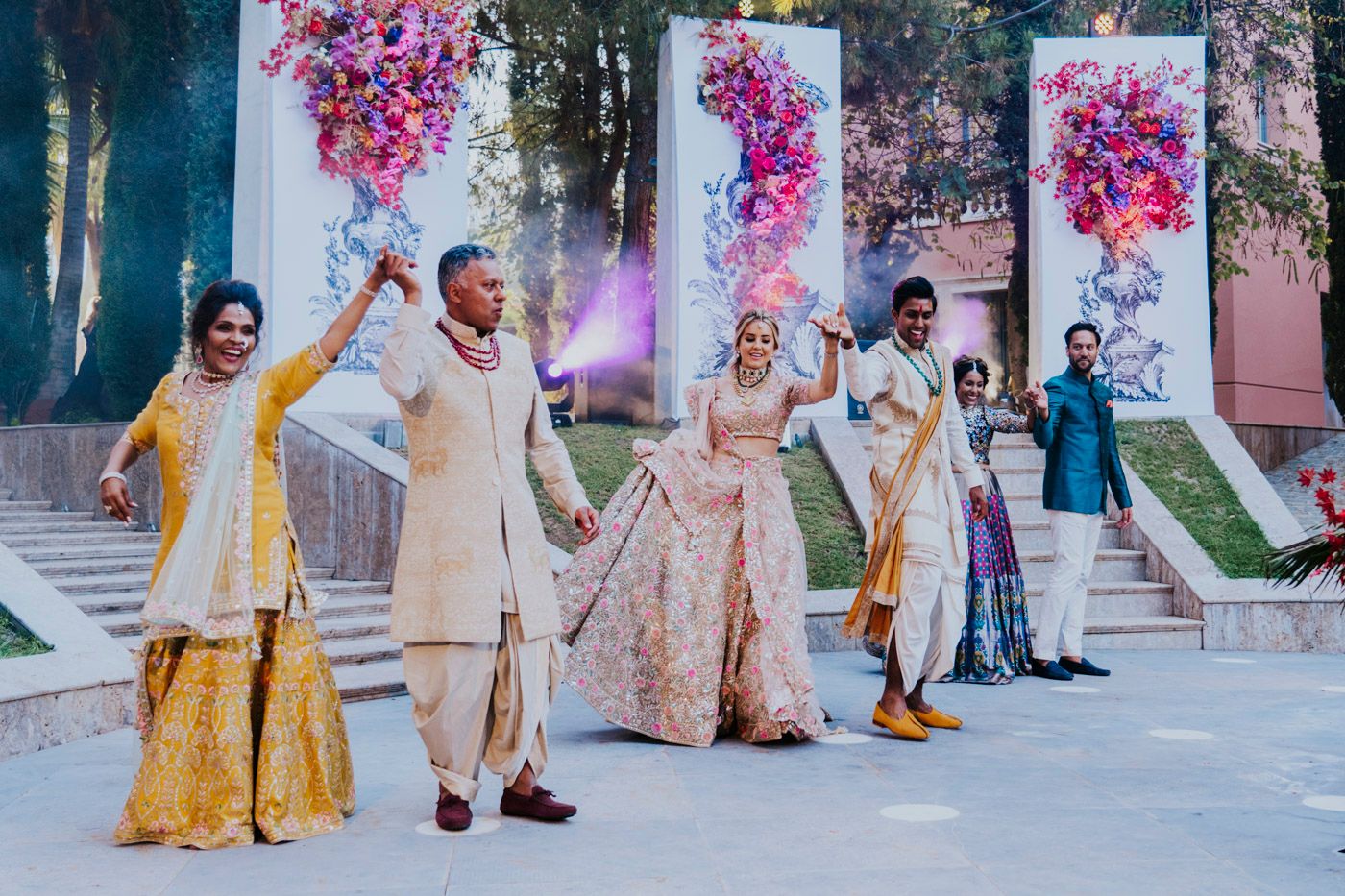 A celebratory group performs a dance sequence with colorful pink and purple smoke effects during an outdoor wedding celebration.