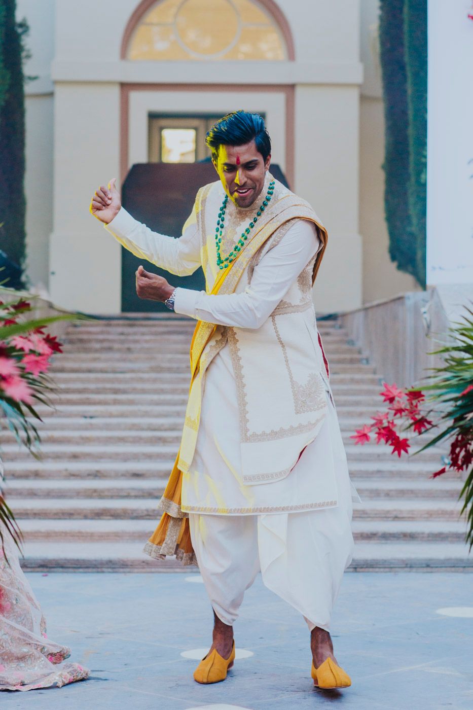 A festive dancer in traditional white Indian attire with yellow shoes performs celebratory moves at an outdoor venue.