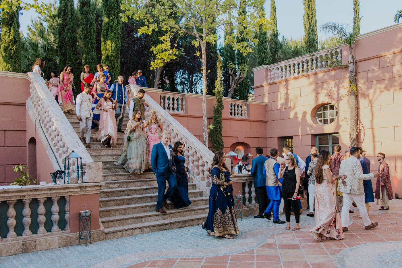 Wedding guests gather on elegant pink marble stairs and balustrades during an outdoor reception event.