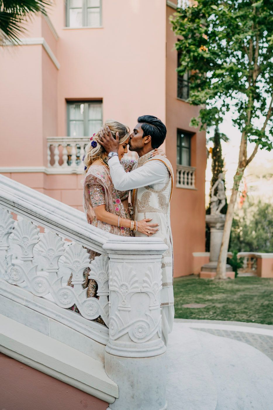 Couple sharing a romantic moment on ornate white marble stairs against a pink stucco building.