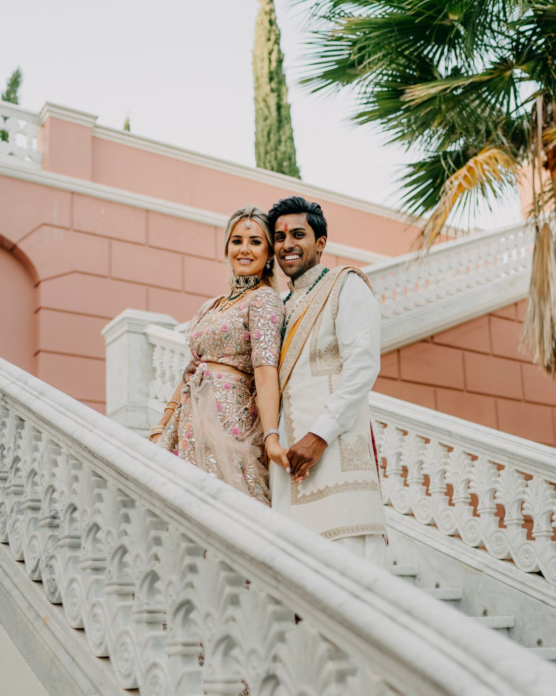 People in elegant attire pose on ornate white marble steps against a coral-colored wall and tropical palm trees.