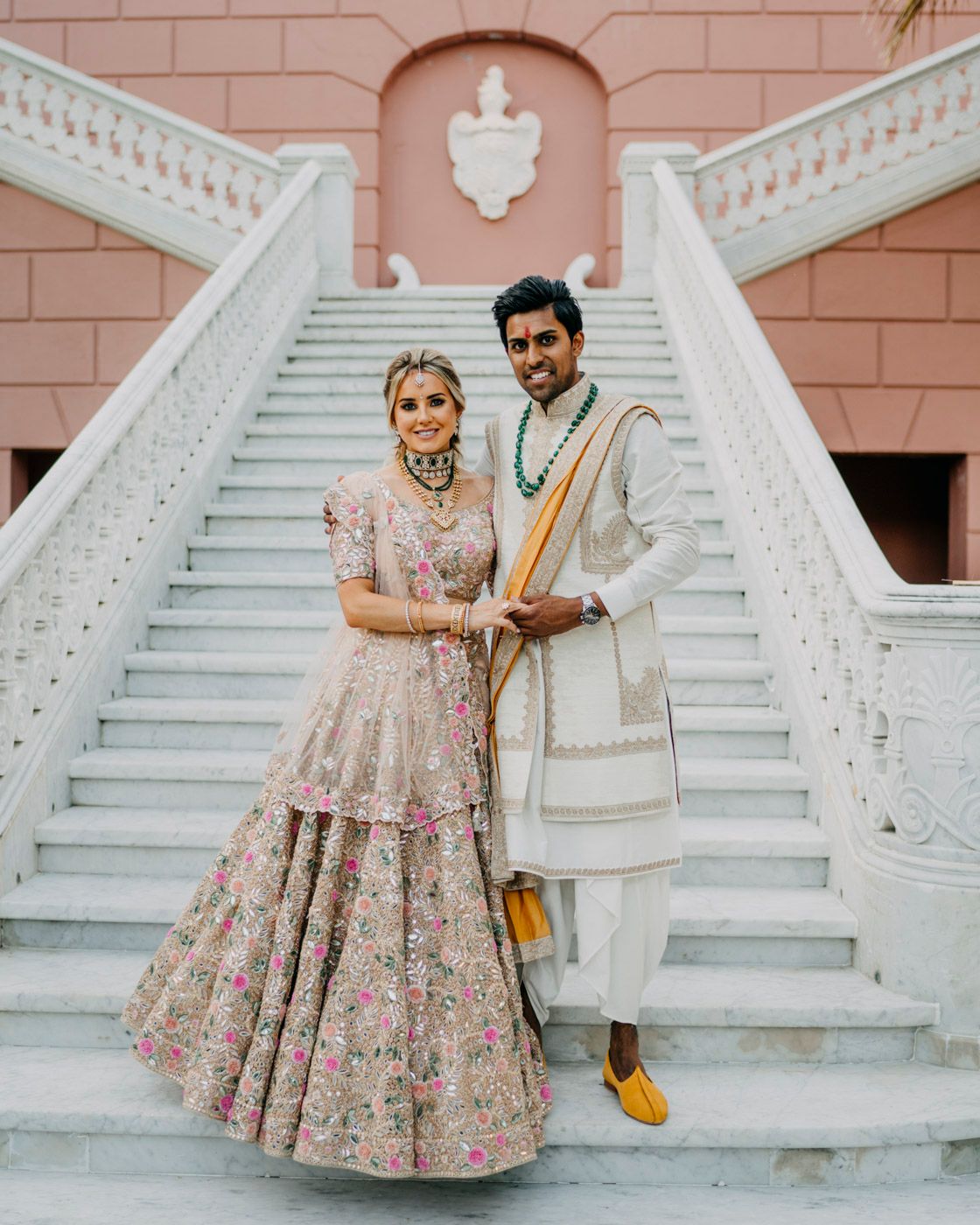 Traditional Indian wedding attire showcased on a grand white staircase against a coral-colored wall.