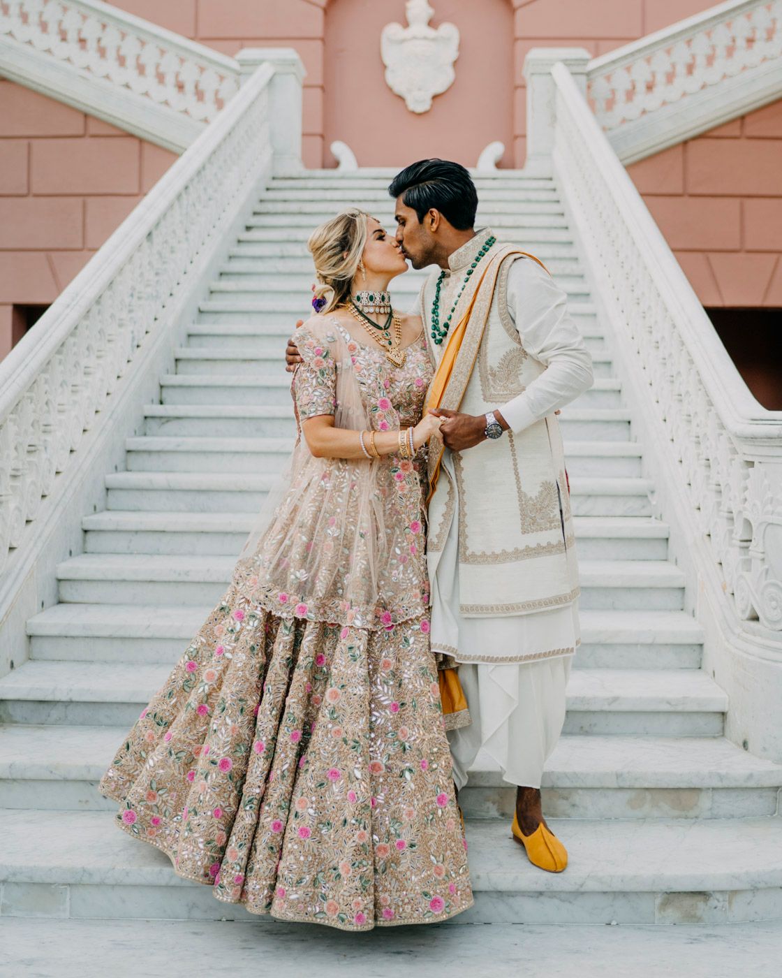 A couple in traditional South Asian wedding attire share an intimate moment on elegant white marble stairs.
