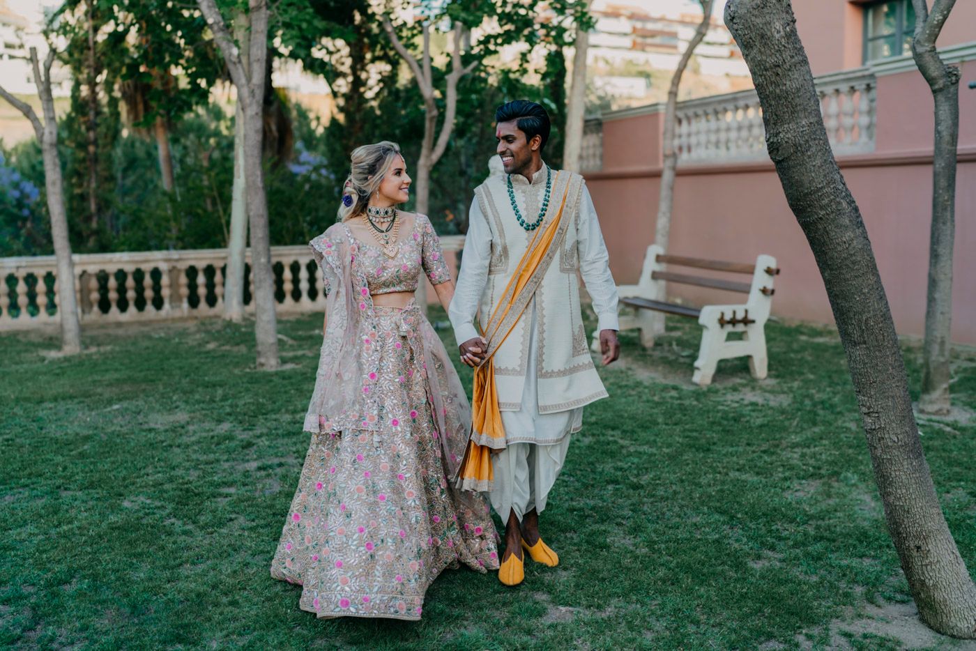 A couple in traditional Indian wedding attire walks together on a lush green lawn beside a pink building.