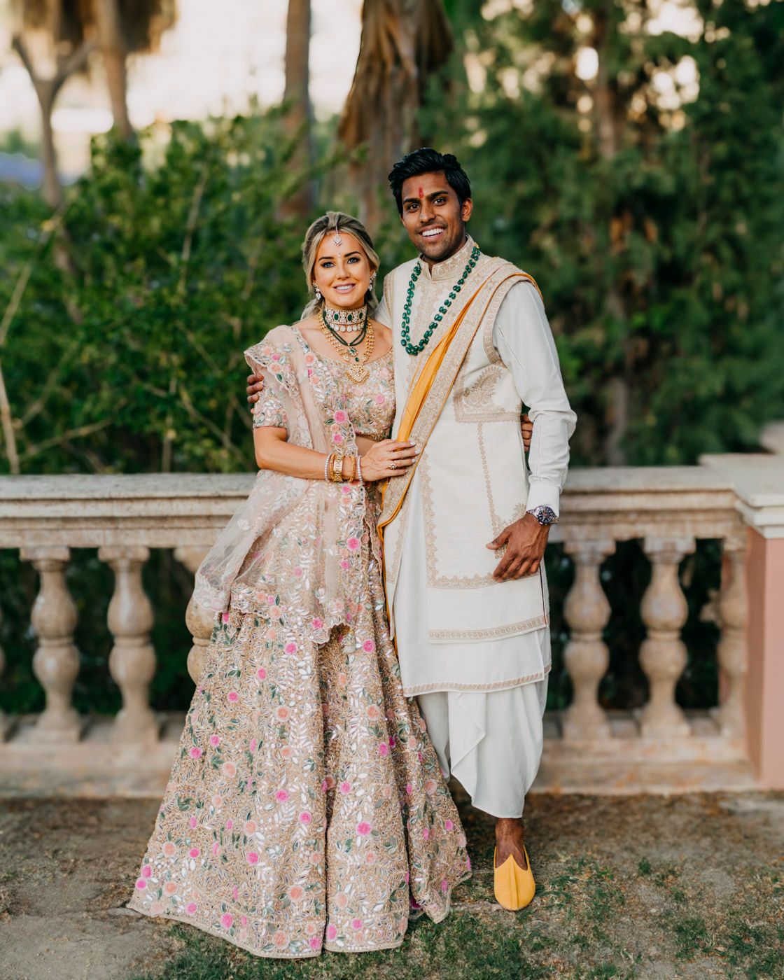 A couple in traditional Indian wedding attire pose together on an outdoor balcony at sunset wearing pink and white outfits.