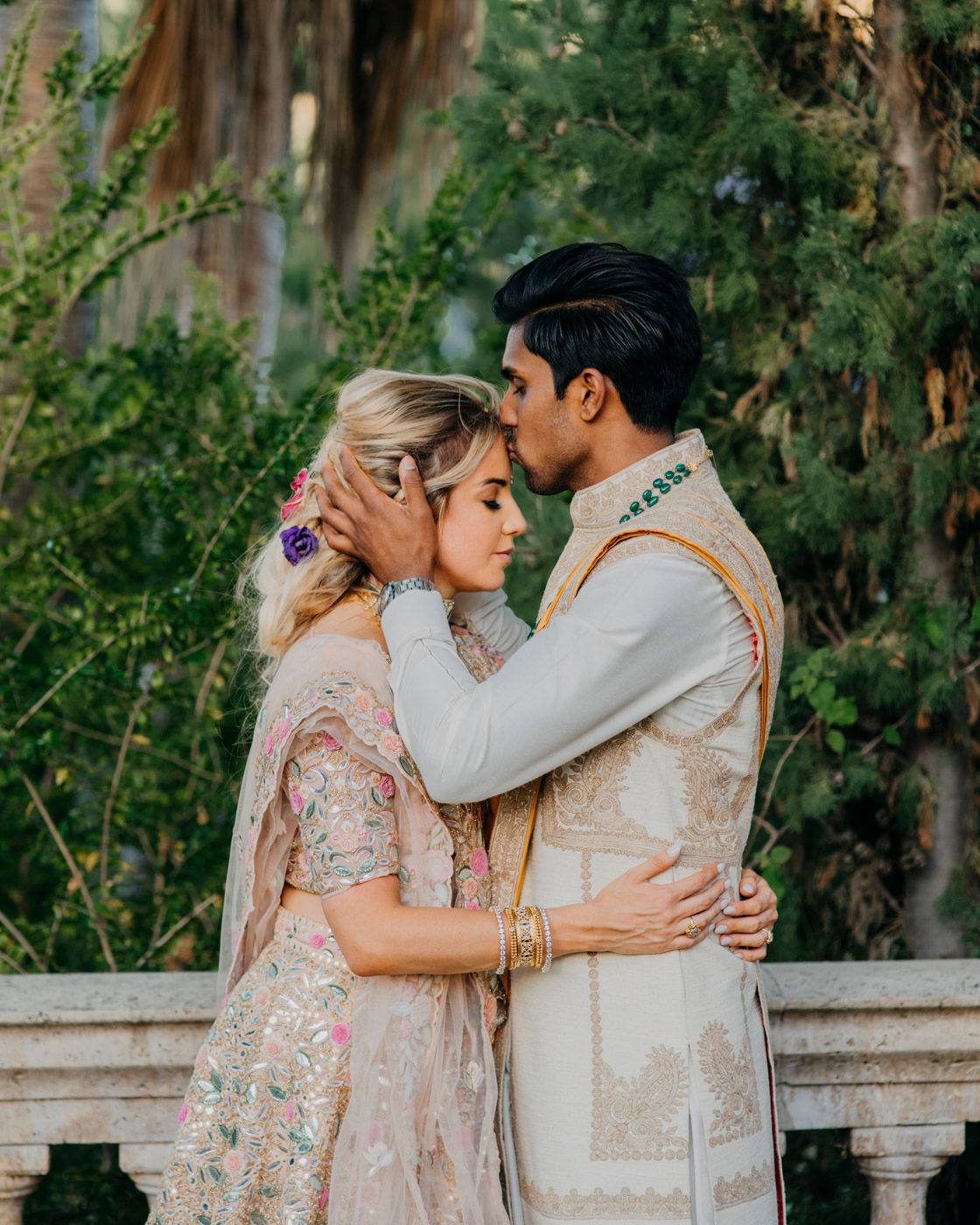 A couple in traditional South Asian wedding attire embrace intimately among trees and greenery during a romantic outdoor moment.