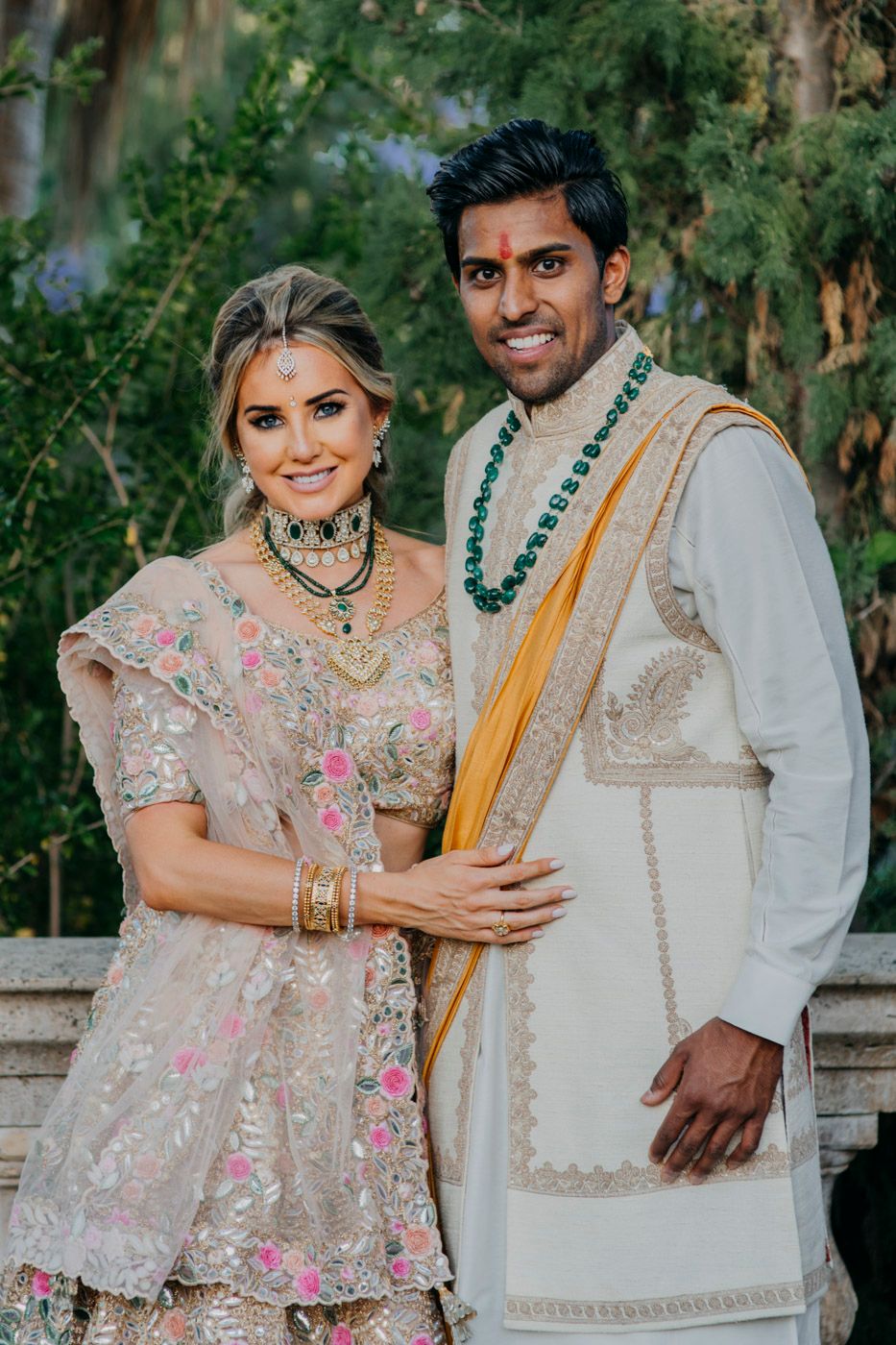A couple in traditional South Asian wedding attire smiling together, wearing ornate embroidered outfits and jewelry.