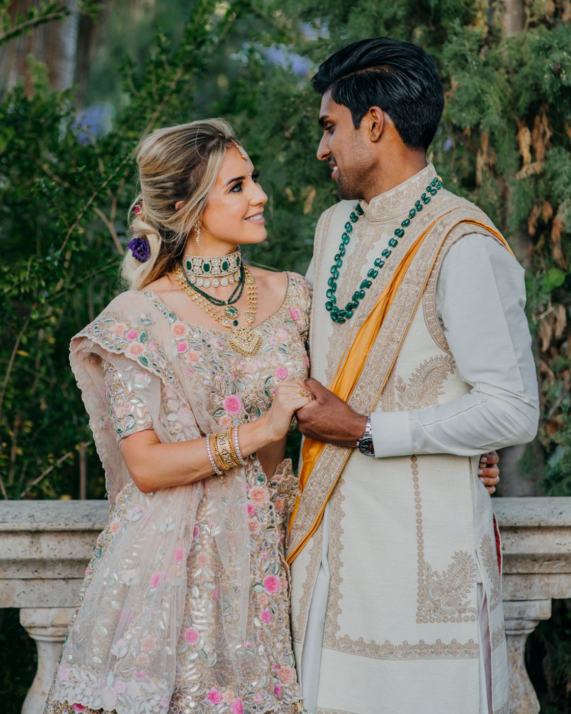 A couple in traditional South Asian wedding attire with embroidered details smile at each other against a leafy backdrop.