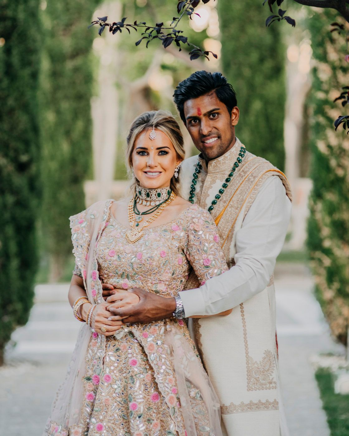 A couple in traditional South Asian wedding attire pose together outdoors among green trees and pillars.