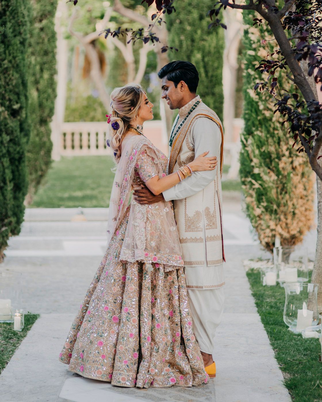 A stunning couple in traditional South Asian wedding attire embrace on a garden pathway lined with candles and cypress trees.