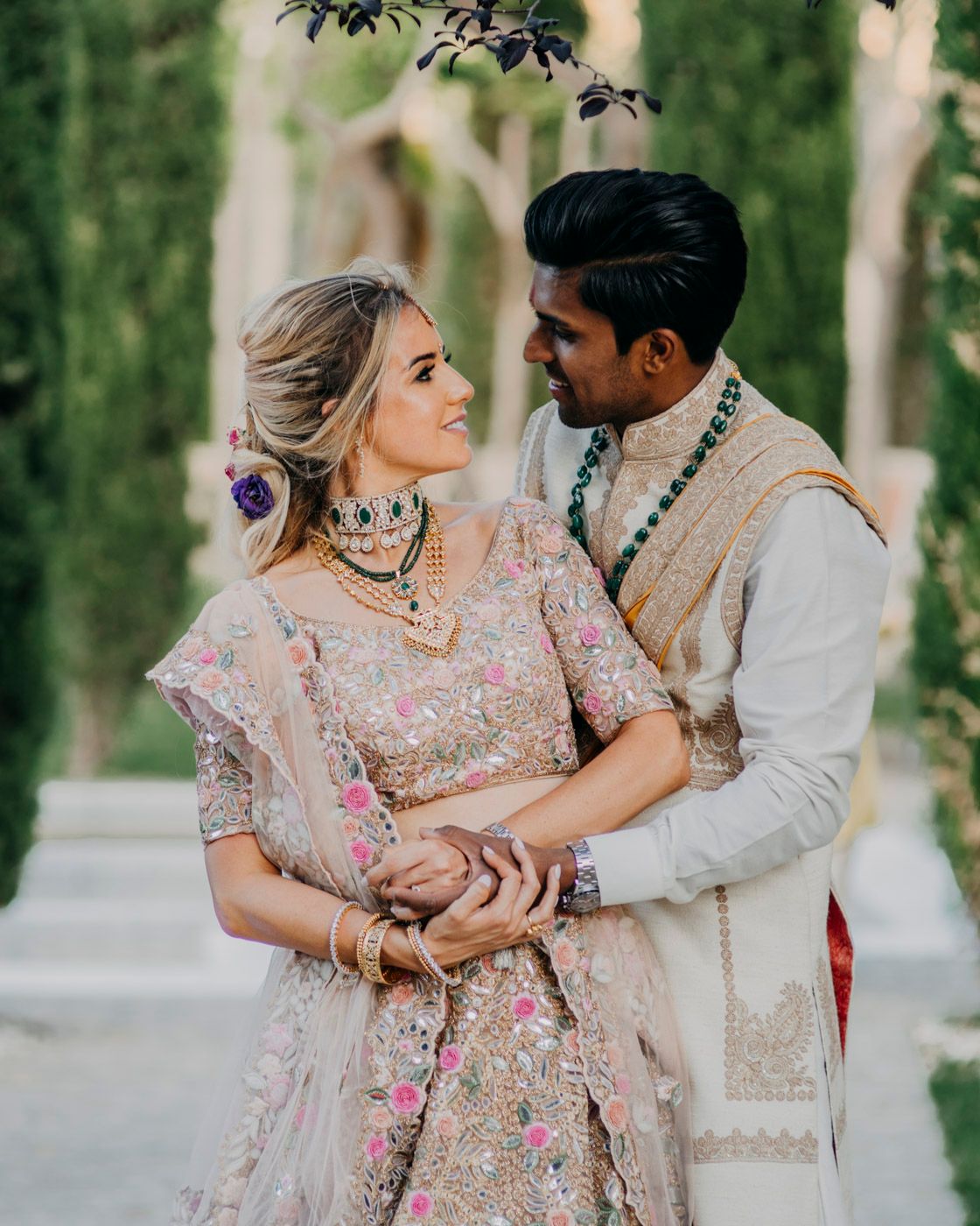 A romantic couple in traditional South Asian wedding attire embrace outdoors among trees in soft natural lighting.