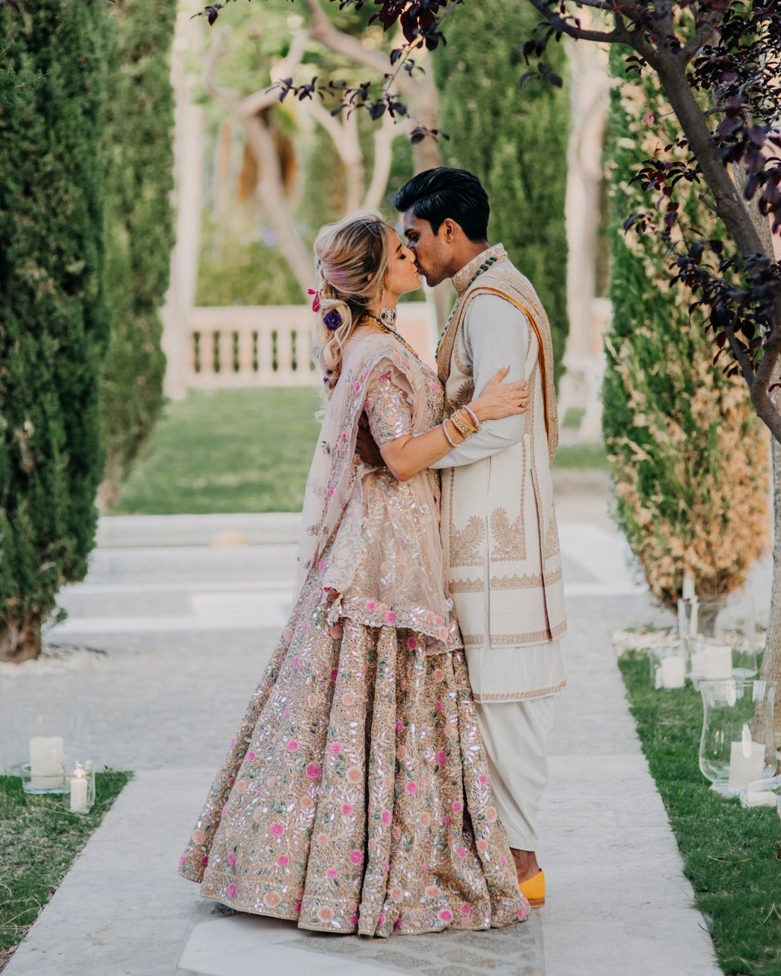 A couple in ornate traditional South Asian wedding attire embrace on a garden path lined with cypress trees and candles.