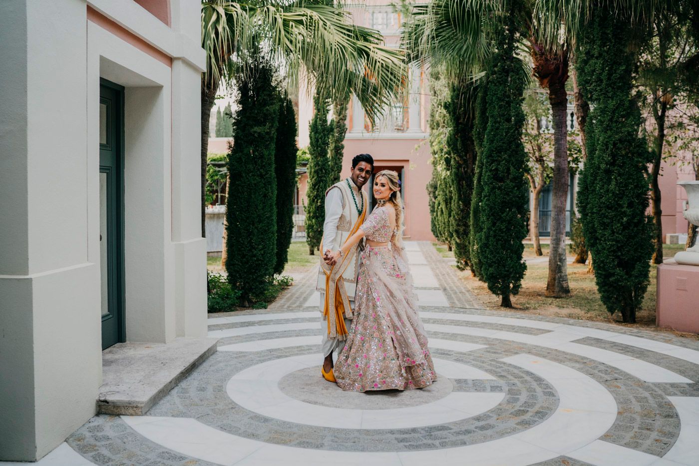 A couple poses in an elegant courtyard with towering cypress trees and a patterned stone floor design.
