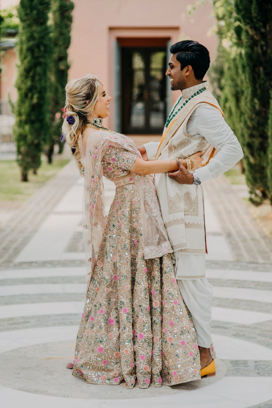 A couple in traditional South Asian wedding attire dance together on a path lined with cypress trees.