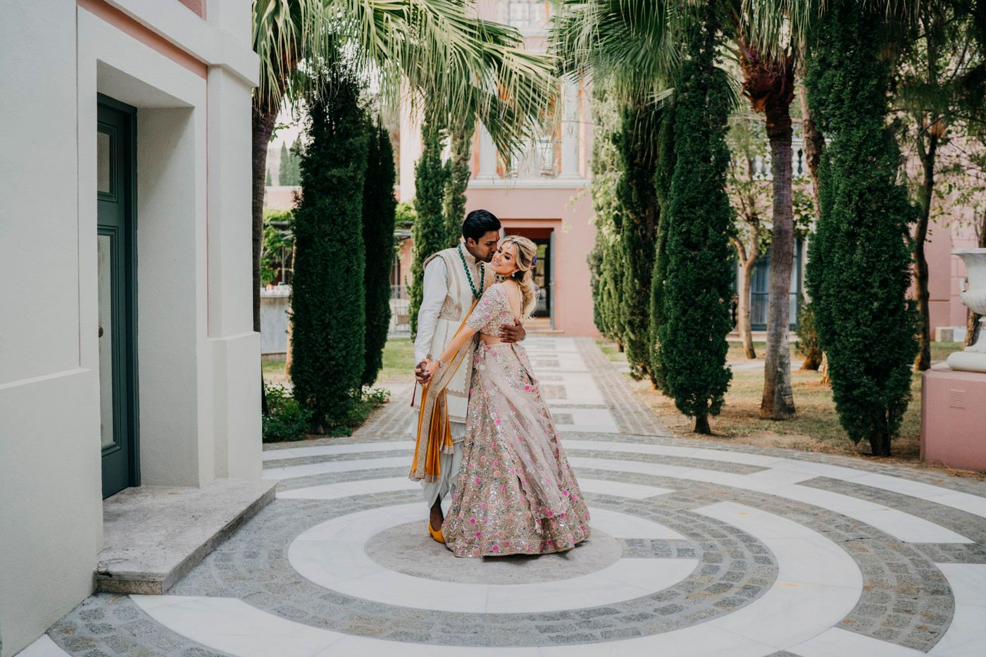 A couple stands together on a circular patterned tile path surrounded by tall cypress trees and pink buildings.