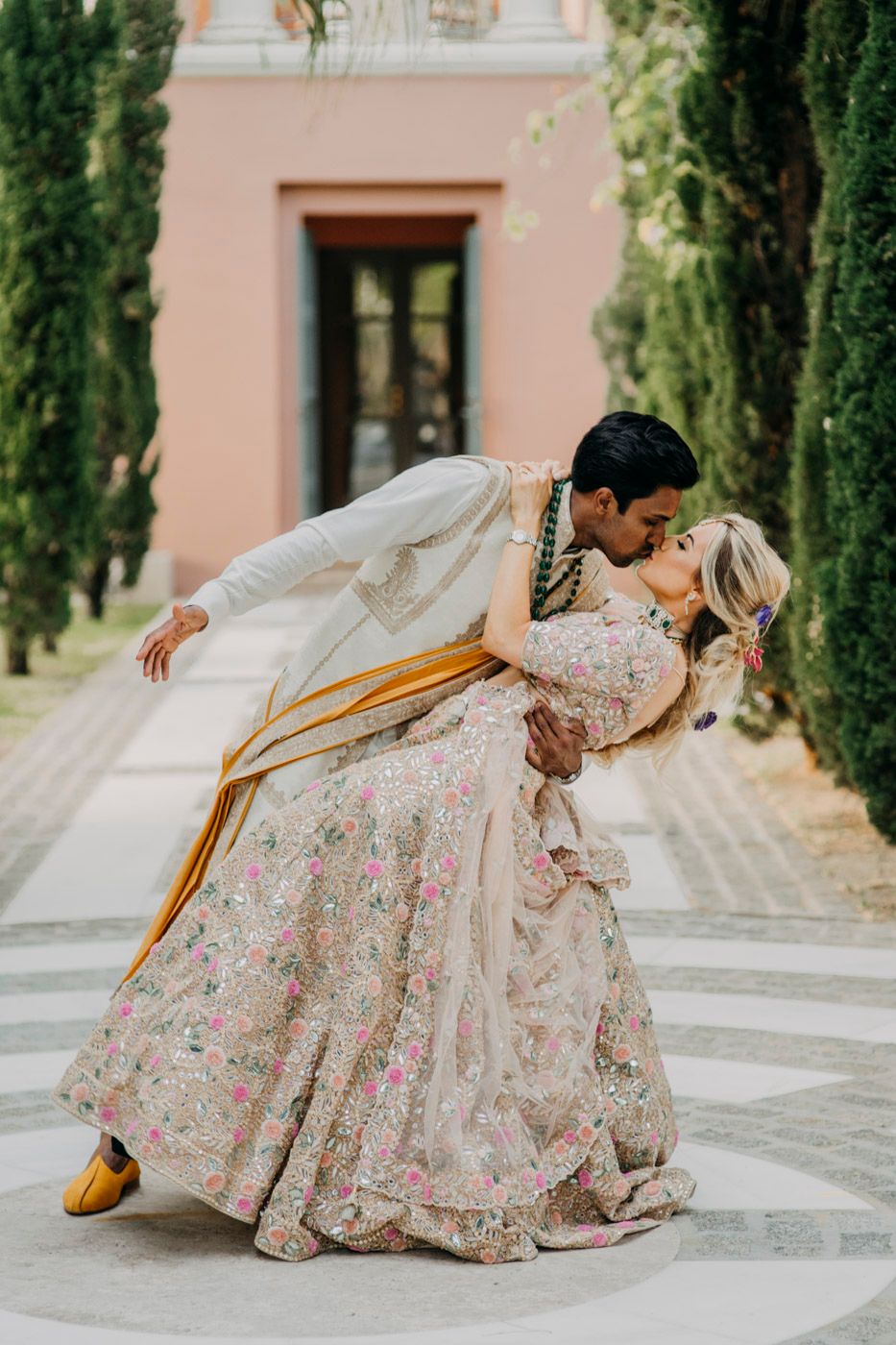 A romantic wedding couple in traditional Indian attire share an intimate dip pose against a pink villa and cypress trees.