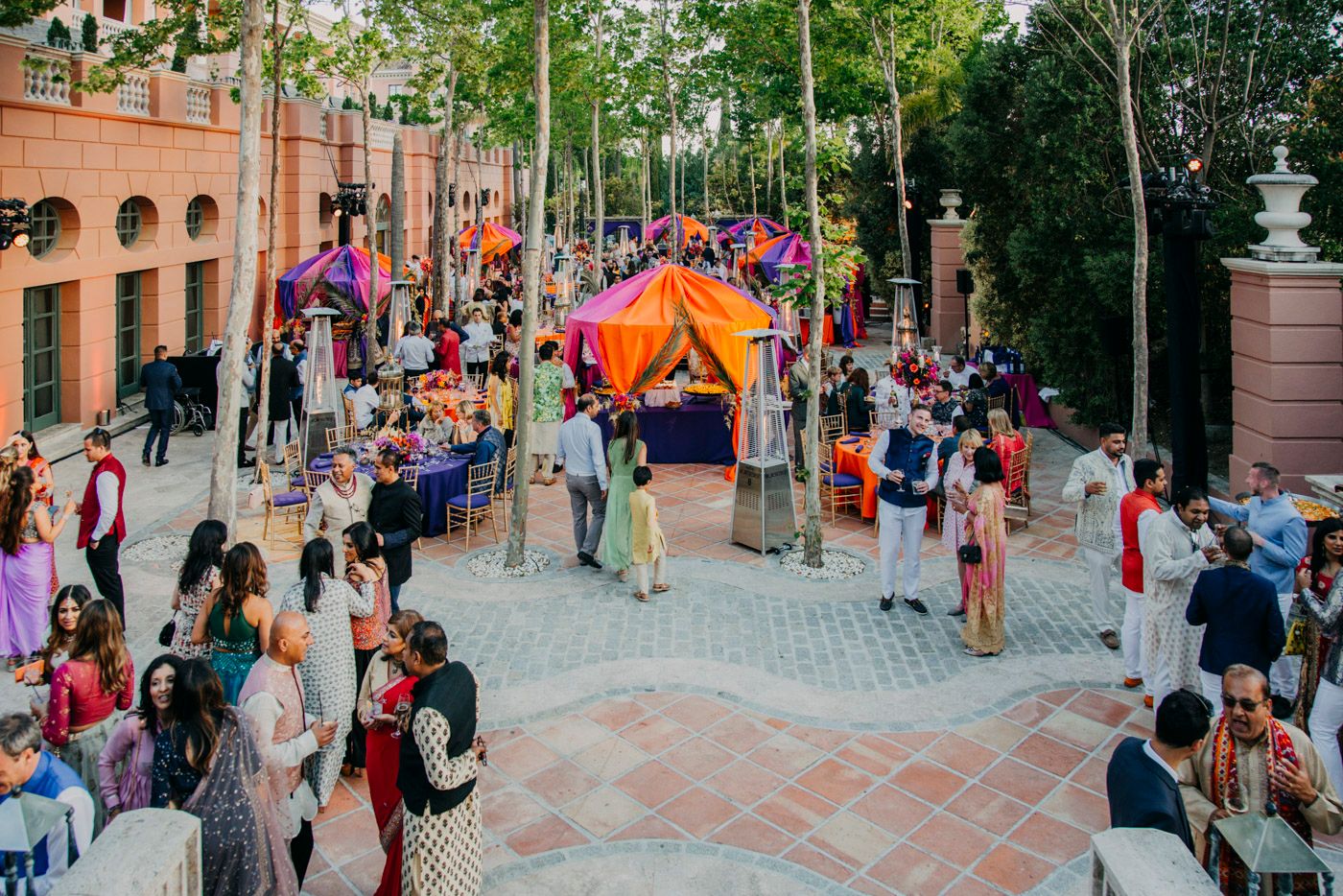 Crowd of people gathering for an outdoor festival with colorful tents and decorations on a brick-paved plaza.