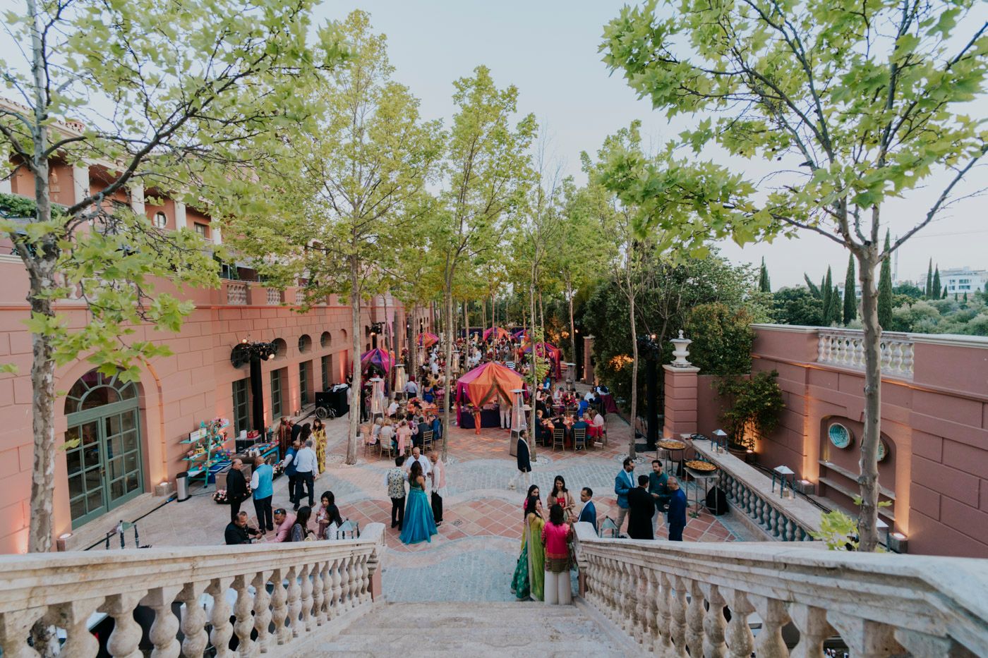 Crowds of visitors gather on a pink-walled Spanish-style plaza with ornate staircases and green trees overhead.