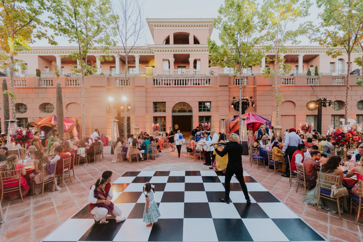 Reception guests dine outdoors at elegant pink venue featuring black and white checkered dance floor and Mediterranean architecture.
