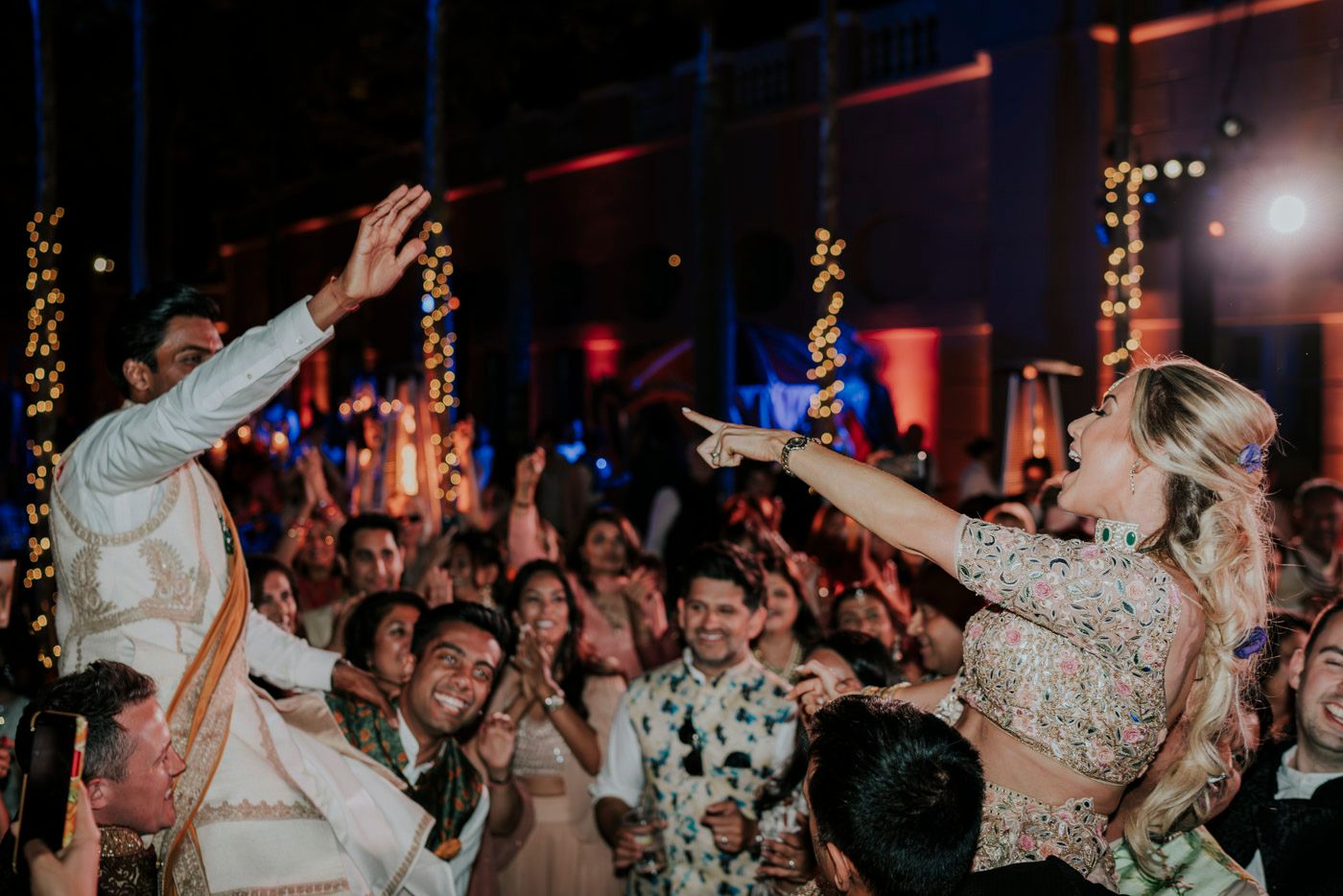 Wedding reception party crowd dancing and celebrating with hands in the air under glowing string lights and red ambiance.