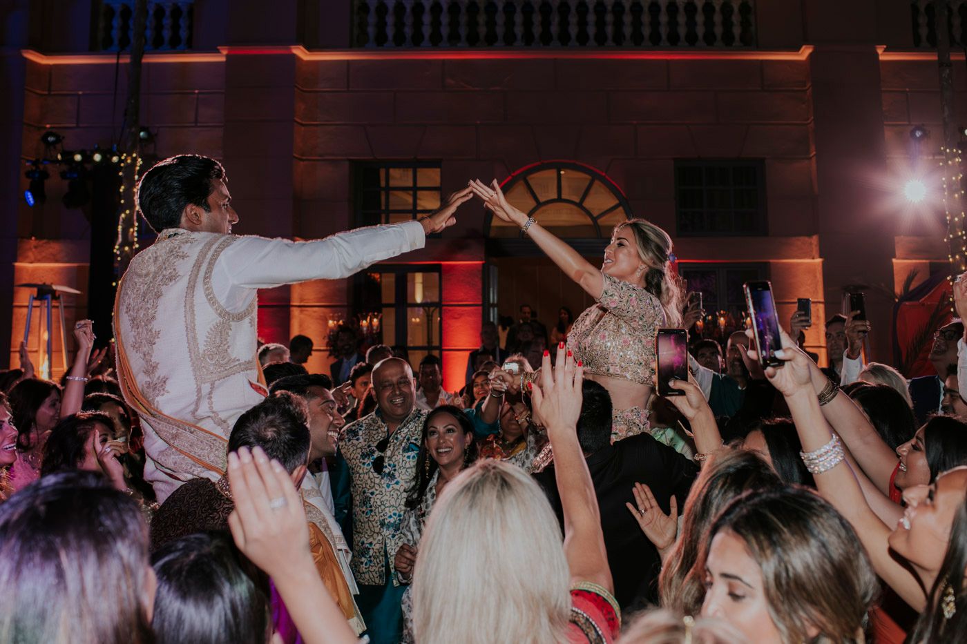 A crowd of people dancing and celebrating at a nighttime wedding reception with vibrant red lighting.