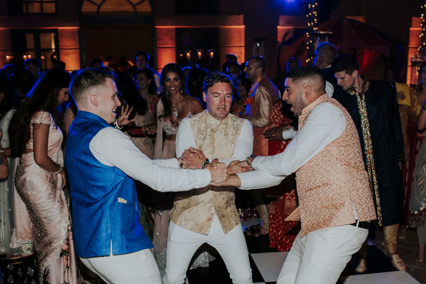 People dancing and celebrating at a festive Indian wedding reception with colorful formal attire and mood lighting.