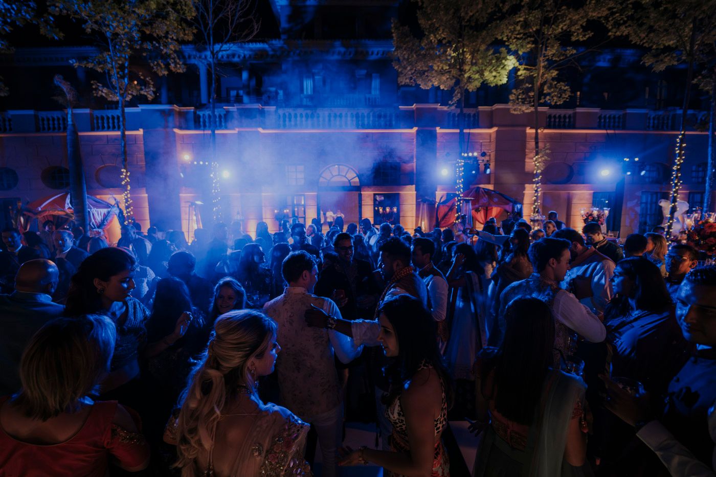 A crowded outdoor party scene illuminated by blue lights and fog with people dancing at night.