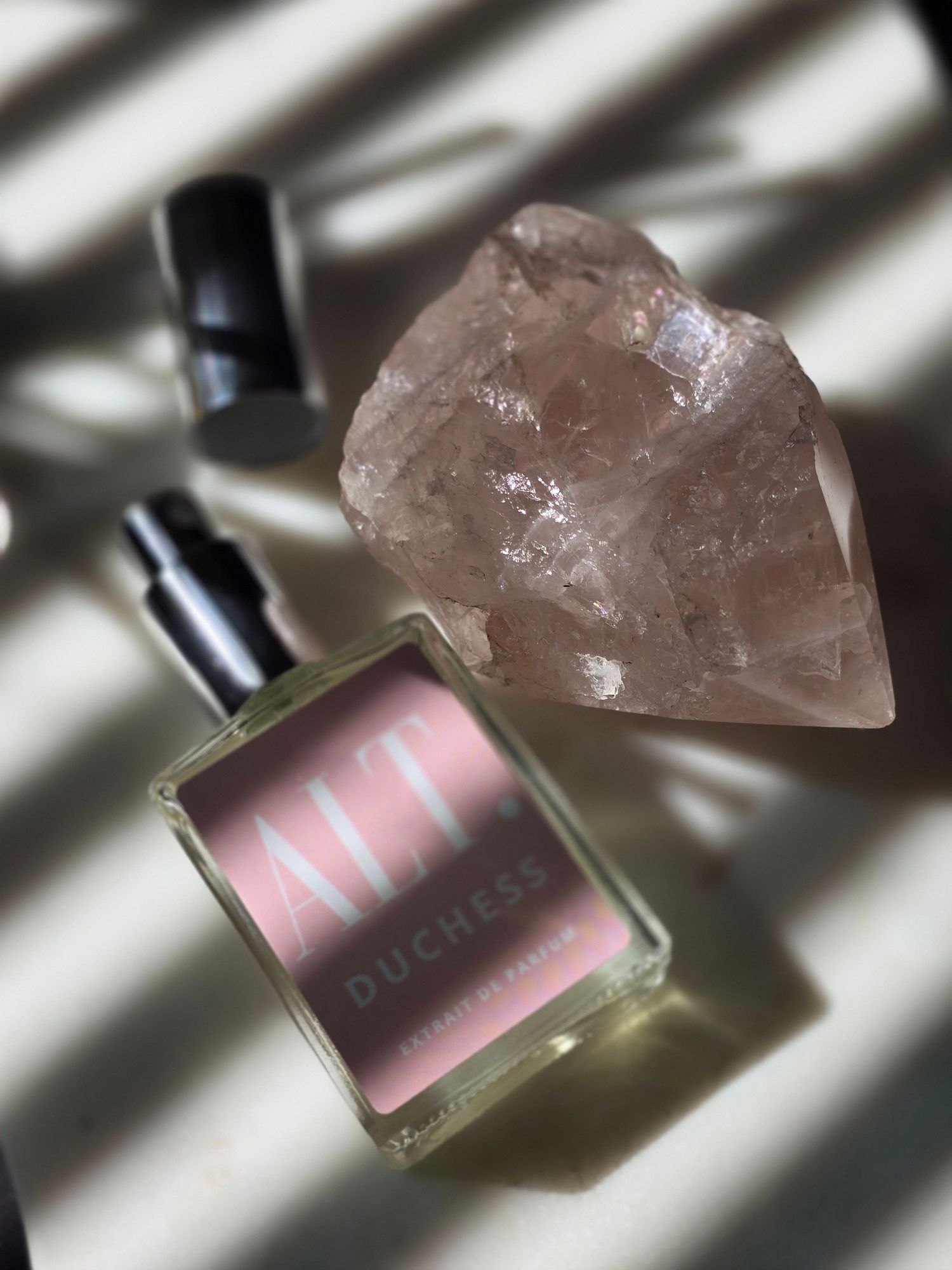 A pink perfume bottle sits beside a raw rose quartz crystal, casting dramatic diagonal shadows.