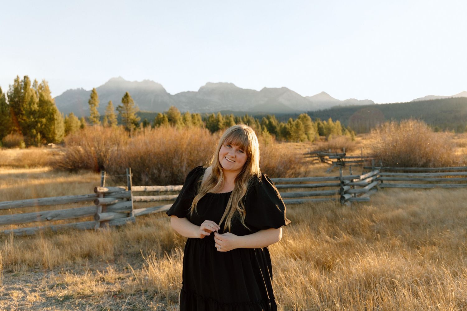A person in a black dress stands in a golden field with mountain ranges and wooden fences in the background at sunset.