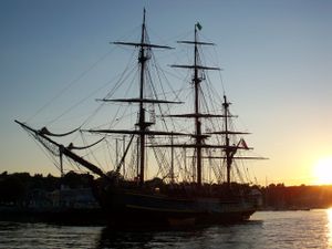 Silhouettes of tall ships at sunset with dramatic sky and water reflections.