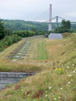 Grassy hillside with rows of memorial markers overlooking a suspension bridge in the distance.