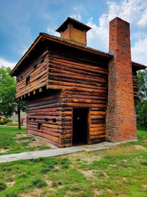 Historic log cabin blockhouse with tall brick chimney stands against bright blue sky.