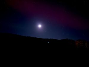 Full moon glows brightly in a dark night sky above silhouetted mountain ridges.